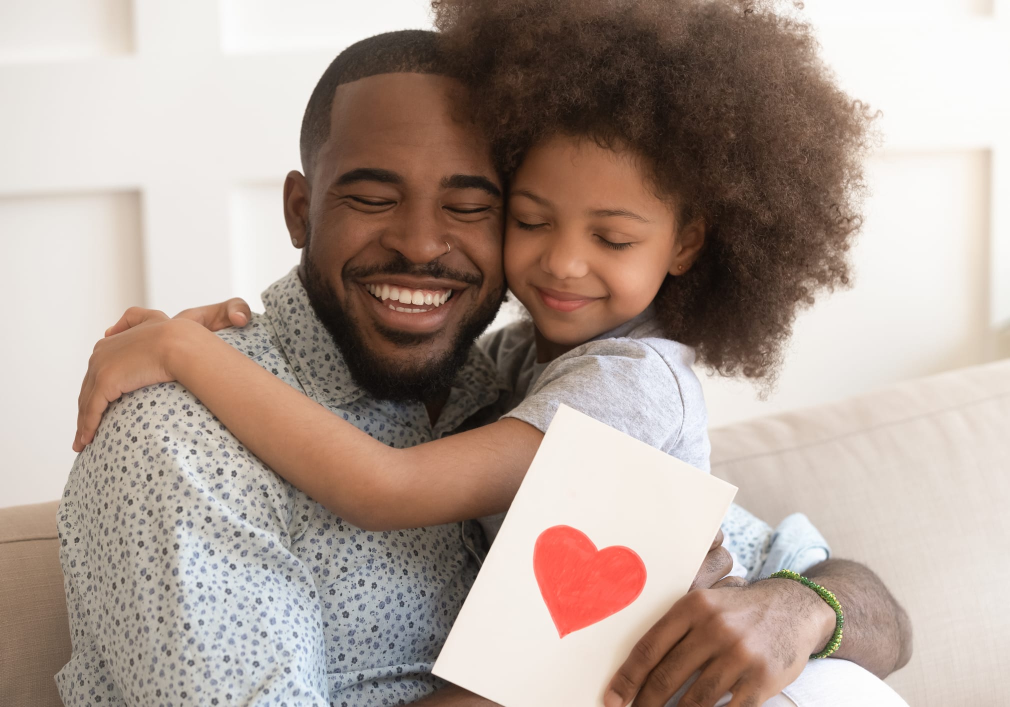 A father embraces his daughter on the couch at home, holding the handmade heart card she made him