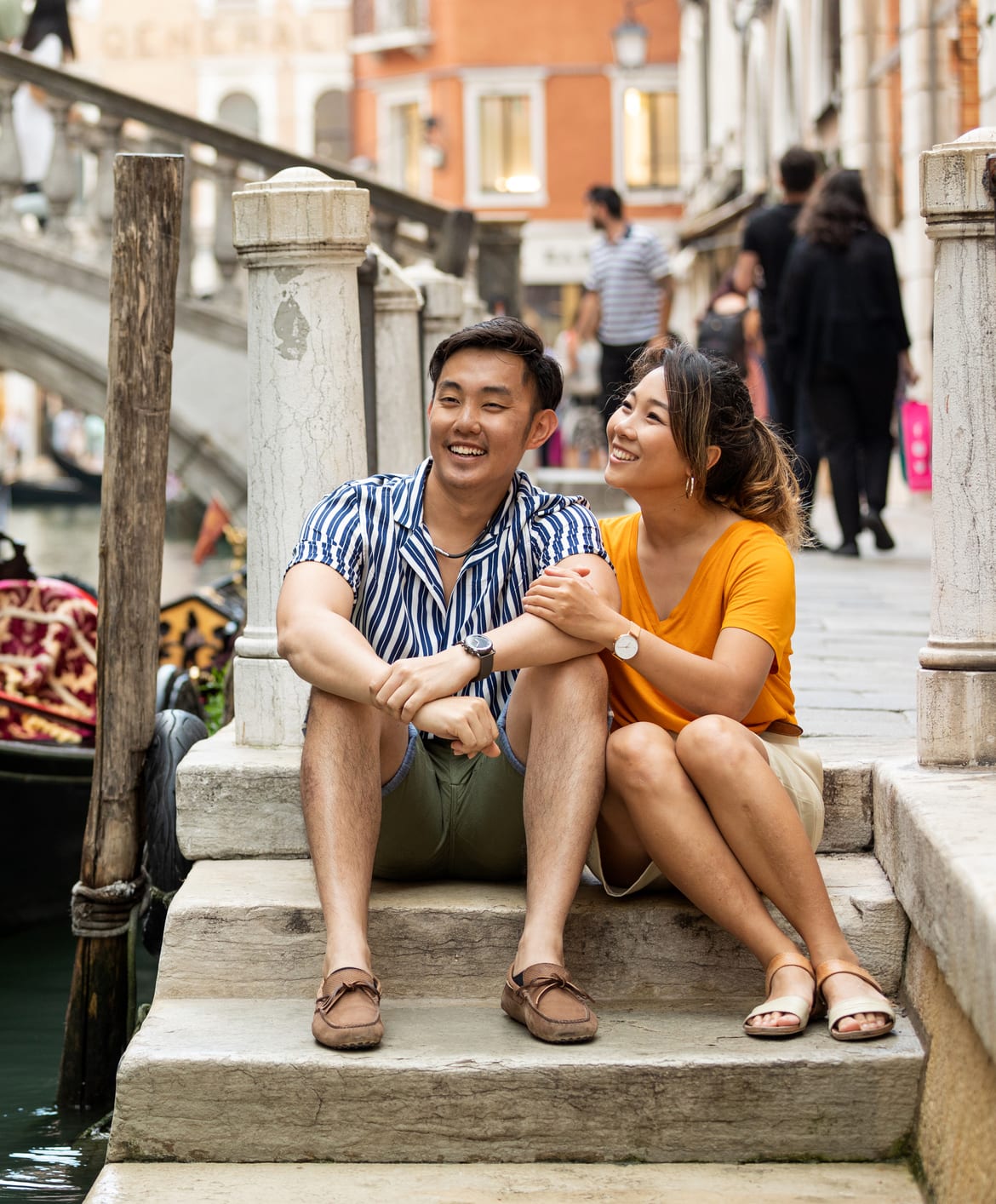 A couple sits together on a stone bridge step above a Venice canal, a gondola moored beside them